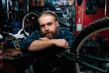 Fototapeta na wymiar Portrait of successful cycling mechanic male standing leaning on bicycle in repair bike workshop with dark interior, looking at camera. Concept of professional repair and maintenance of bicycle.