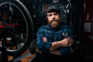 Fototapeta na wymiar Brutal cycling mechanic male in cap holding wrench in hand standing by bicycle in repair workshop with dark interior, looking at camera with serious expression. Concept of bicycle maintenance.
