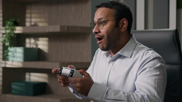 Funny lazy business worker addicted video game Indian man playing console controller play online at workplace office afraid scared get caught Arabian businessman hide joystick pretend working computer