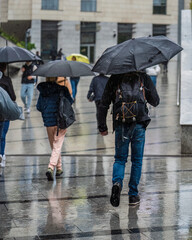 Authentic young people with umbrellas walk in the rain along the city street, rainy day in the city, reflections of soaked passers-by on the wet road, raindrops and puddles. Vertical frame