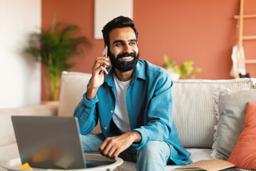 Cheerful indian man with laptop communicating on phone at home