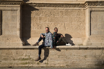 Real marriage of gay couple, sitting on a stone wall with backs together and holding hands, looking at camera smiling and happy. Concept lgtb, lgtbiq+, couples, in love, love.