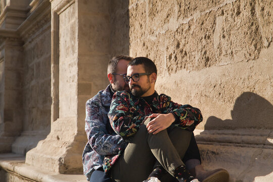Real marriage of gay couple, sitting on a stone wall while one embraces the other from behind with very affectionate, complicit and happy attitude. Concept lgtb, lgtbiq+, couples, in love, love.