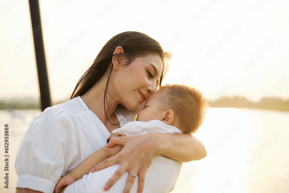 Wall mural portrait of happy mother travel in park on weekend with sunset view, mother holding baby son relaxin