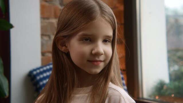 Portrait of a cute little girl posing indoors. Stock footage. Beautiful smiling girl touching her long hair and posing by the window.