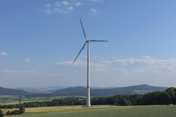 Fototapeta premium Trendelburg, Germany - 06/13/2023: Single wind turbine on the Deiselberg in a rural landscape with fields, meadows, trees, forests, mountains and valleys with a blue sky with little cloud cover.