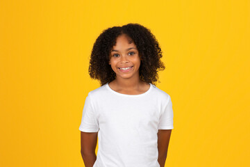 Cheerful curly teenager black schoolgirl in white t-shirt looking at camera, isolated on yellow...