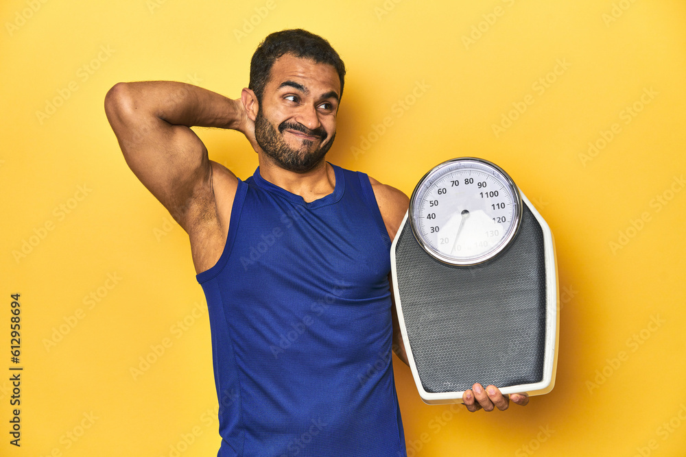 Wall mural Fit young Latino man with scale on yellow background, fitness concept, touching back of head, thinking and making a choice.