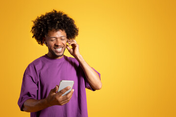 Cheerful black adult curly man in purple t-shirt and wireless headphones looks at phone - obrazy, fototapety, plakaty