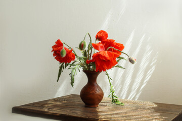 Summer bouquets: a bouquet of red poppies in a brown vase on a wooden table, light background, sun rays, space for text
