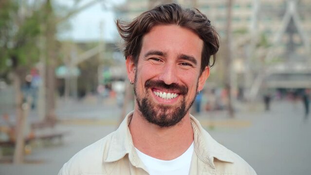 Close up portrait of handsome bearded guy with perfect white teeth smiling and looking at camera standing outdoors. Front view of young adult man with friendly and positive expression. Happy male