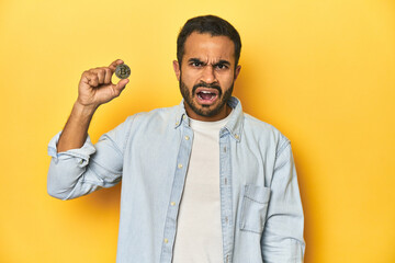 Young Latino man holding a Bitcoin coin, yellow studio background, screaming very angry and aggressive.