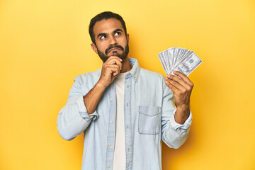 Young Latino man holding a bundle of dollars, yellow studio background, looking sideways with...