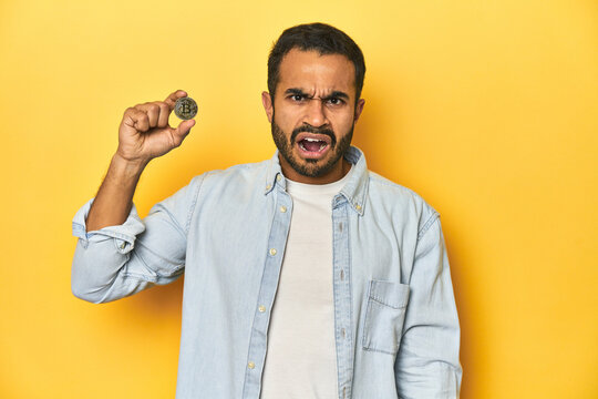 Young Latino man holding a Bitcoin coin, yellow studio background, screaming very angry and aggressive.