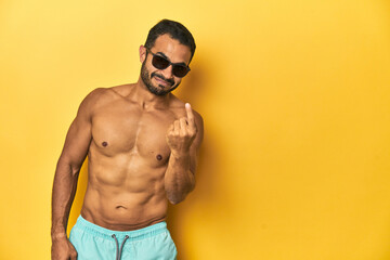 Fit young Latino man in swimwear and sunglasses, yellow studio background, pointing with finger at you as if inviting come closer.