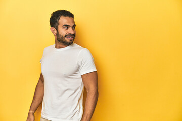Casual young Latino man against a vibrant yellow studio background, looks aside smiling, cheerful and pleasant.