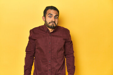 Casual young Latino man against a vibrant yellow studio background, shrugs shoulders and open eyes confused.
