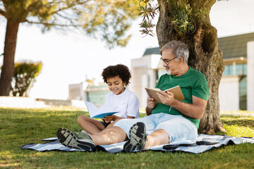 Cheerful senior man with beard and black little boy sit on plaid, read book, relax in park, outdoor