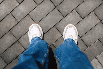 Women's legs in jeans and white modern sneakers on gray street tiles, copy space, top view, personal point of view