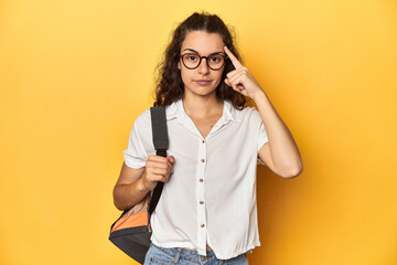 Caucasian university student with glasses, backpack, pointing temple with finger, thinking, focused on a task.