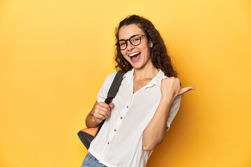 Caucasian university student with glasses, backpack, points with thumb finger away, laughing and...