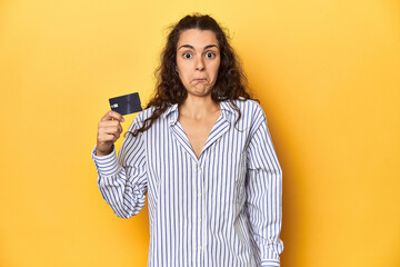 Woman holding a credit card, yellow backdrop, shrugs shoulders and open eyes confused.