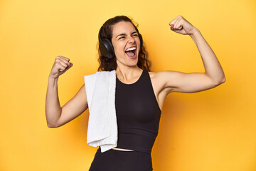 Athletic woman holding a towel, yellow background, raising fist after a victory, winner concept.