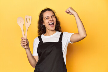 Woman with apron, wooden cooking utensils, yellow, raising fist after a victory, winner concept.