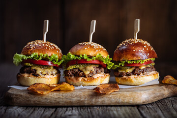 Three burger sliders placed on a wooden board in rustic atmosphere. Very tasty burgers with melted cheese, lettuce, tomato and sauce - obrazy, fototapety, plakaty