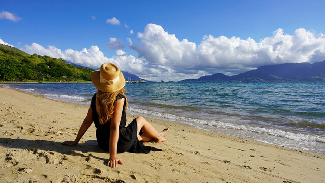 Attractive young woman with straw hat sitting on sand beach in her summer holidays. Copy space.