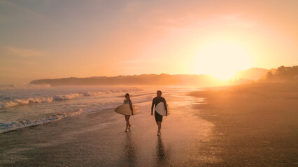 Fototapeta na wymiar AERIAL: Cheerful surfers admire big waves while walking on sandy shore at sunset