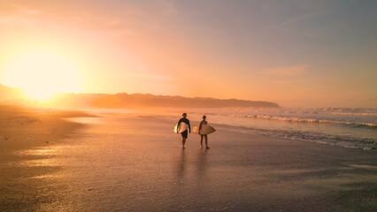 Fototapeta na wymiar AERIAL: Young couple walking on beautiful sandy beach after sunset surf session