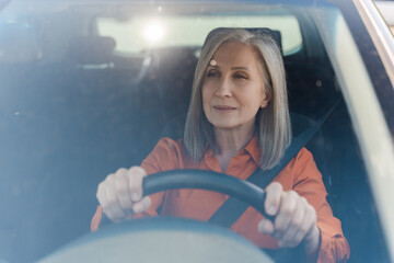 Mature gray haired woman driver holding hands on steering wheel, driving car with fasten belt.