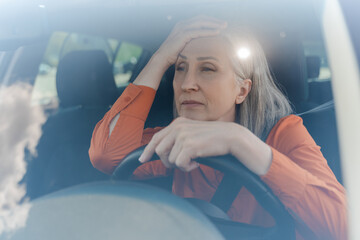 Exhausted mature adult woman driving a car with a fastened seat belt. Traffic jam. Road insurance.