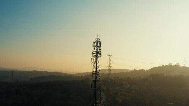 4K drone shot of telephone mast of cell tower. Aerial view of telecom antenna and cellular base station against sunrise and small town surrounded by forests and hills during golden hour stock video.