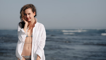 Portrait of a European girl in a white shirt and bikini, on the shore of Bali beach, smiling, fooling around. Have fun on the ocean coast. Freedom, rest in tropical countries.