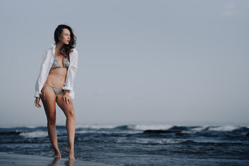 Young girl in bikini and white shirt playing with sea waves on the shore of a Balinese beach. Travel to tropical countries.