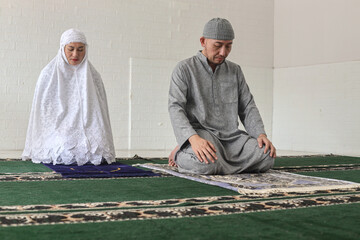 Asian Muslim man becoming imam and leading his wife to pray, doing one of movement gesture in salat procedure. The worshiper proceeds to sit and recite prayers