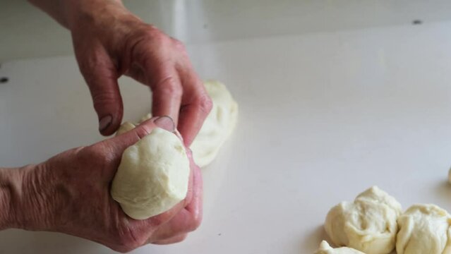 Close-up of wrinkled hands of an elderly woman pinching off raw dough to form balls. An old grandmother bakes buns using a traditional family recipe.