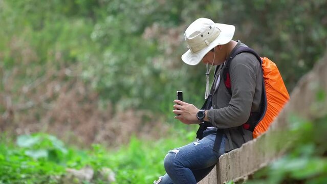 A young man who enjoys trekking for a leisure holiday rests. Pause at the edge of a small stream and bring cell phone to take pictures of the surrounding natural beauty of the forest.