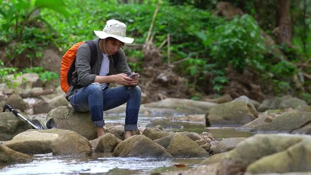 A young man who enjoys trekking for a leisure holiday rests. Stop to sit and rest by the stream to recover from exhaustion. Exciting adventure and trekking through beautiful nature.