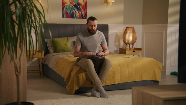 Concentrated man sits on the bed in bedroom and plays on traditional djembe drum. Musician practices skills in cozy home. House with minimalist modern design. Concept of music training. Static shot.