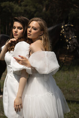 Two beautiful girls in wedding dresses stand next to a horse in the forest