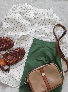 A set of women's comfortable clothes for city walks - cotton bermuda shorts, braided leather sandals, muslin blouse and cross body bag on a light background, top view