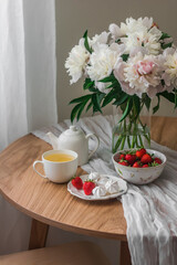 Cozy summer tea party - chamomile tea, meringue, fresh strawberries on a wooden round table with a bouquet of peonies in a glass vase