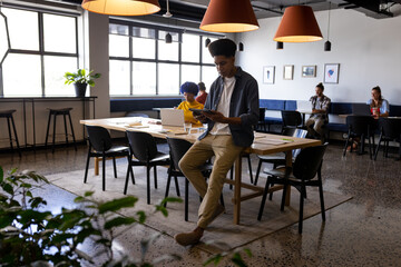 Fototapeta na wymiar Biracial male creative sitting on desk using tablet in casual office
