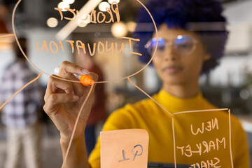 Biracial casual businesswoman with blue afro making notes on glass wall in office