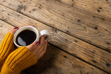 Fototapeta na wymiar High angle cropped hands of caucasian woman wearing yellow sweater holding coffee cup on table