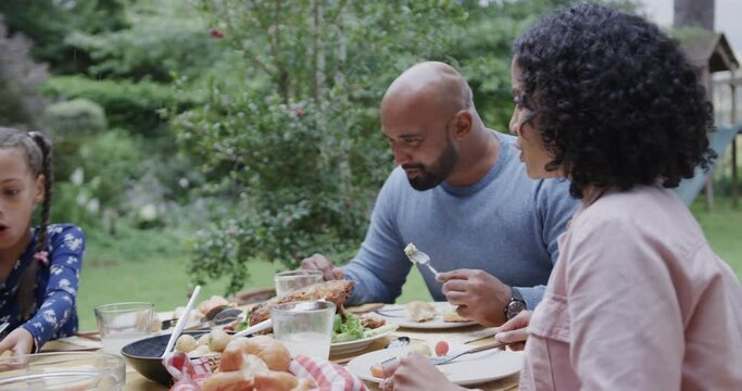Happy biracial parents and daughter eating meal at dinner table in garden, slow motion