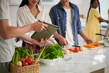 Young man showing online recipe on tablet screen to his wife standing by kitchen table and preparing vegetable salad or other dish with friends